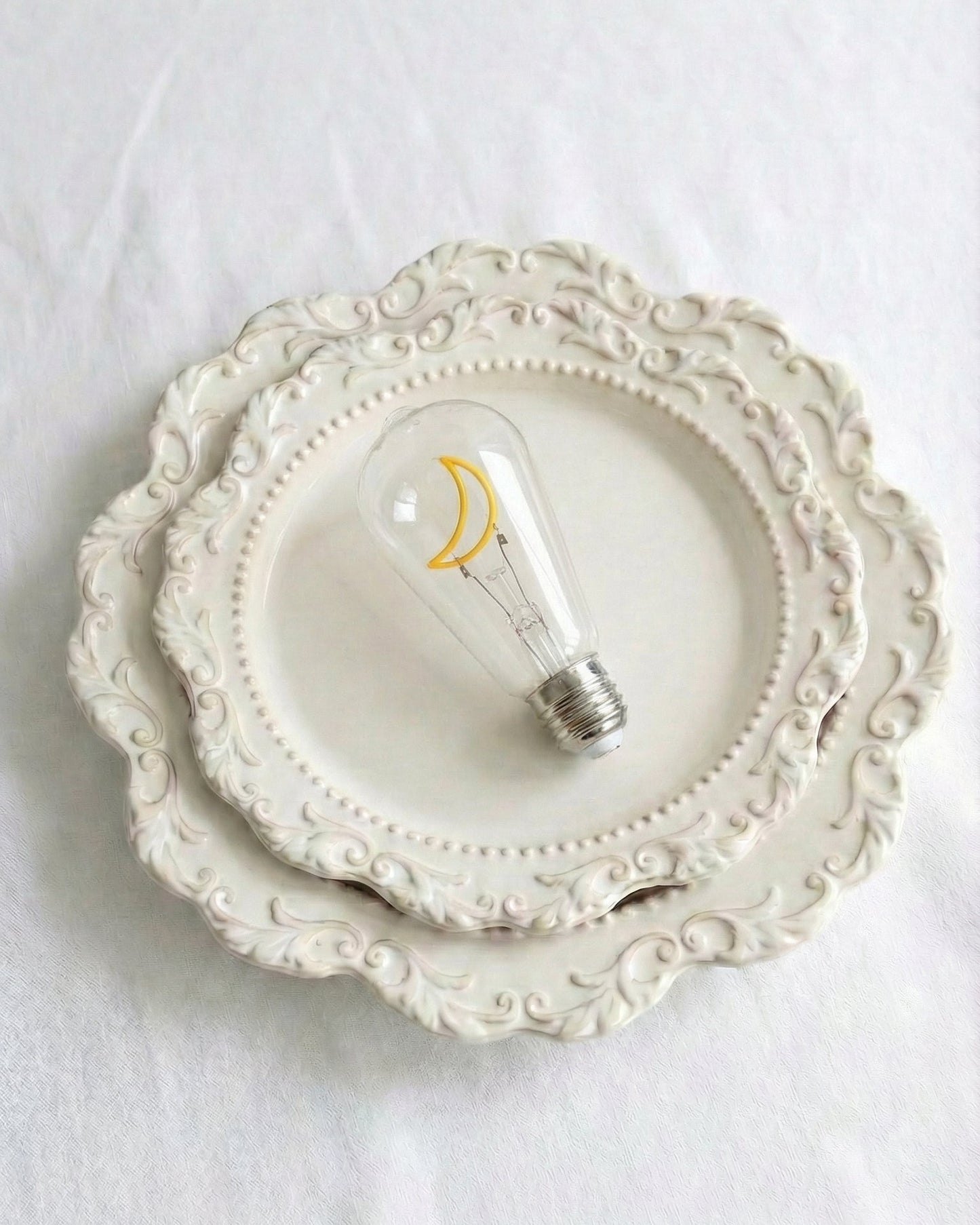 A detailed close-up photograph of a clear glass specialized replacement decorative bulb for a Chavanc_Araromatherapy diffuser, featuring an intricate, non-lighting, yellow moon-shaped decorative element (filament silhouette) inside. The metal connector base is visible at the bottom. The bulb rests on ornate, stacked cream-colored ceramic plates with scalloped edges and beaded borders. Perfect rustic aesthetic accessory for celestial home decor, high-end tablescapes, or home fragrance setup. conversation sta