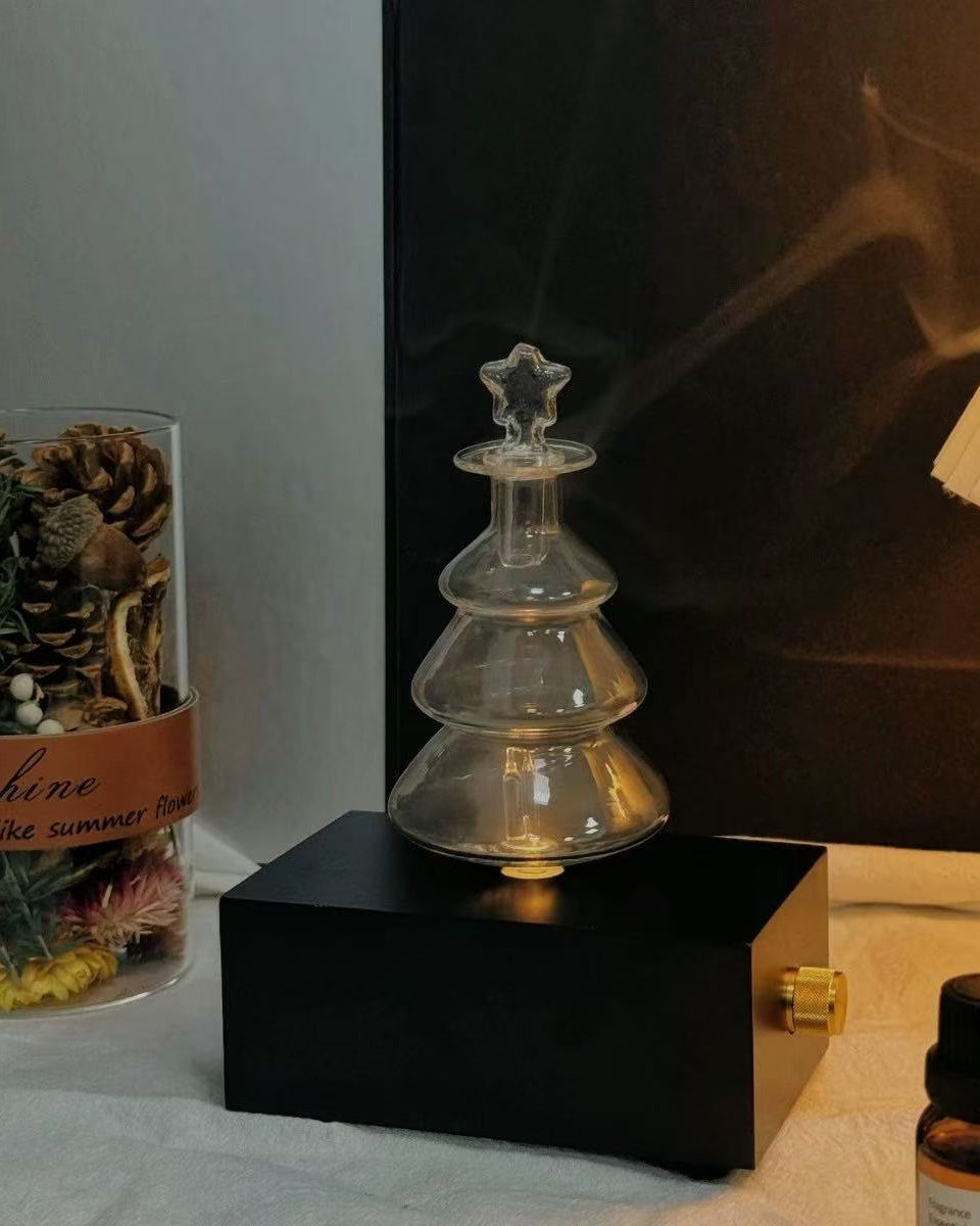 A detailed close-up of a specialized clear glass replacement nebulizing flask accessory for a chavanc aromatherapy diffuser, expertly shaped like a minimalist Christmas tree. The tiered glass flask sits prominently on the same two stacked ornate, cream-colored ceramic plates with scalloped edges and beaded borders as seen in previous images, against a soft white textured fabric background. A fine, cool mist is visibly curling from the tip of the glass tree, simulating its functional use for home fragrance