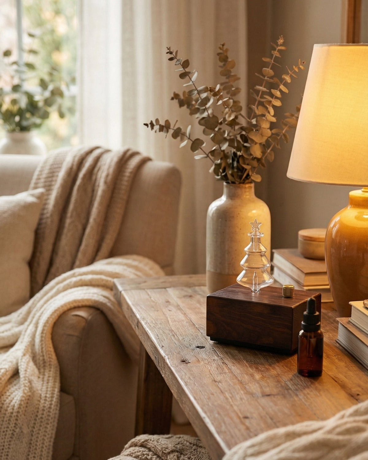 A professional lifestyle photograph of a chavanc nebulizing essential oil diffuser with a dark walnut wood base and a knurled gold knob, topped with a clear glass Christmas tree-shaped flask. The diffuser is placed on a rustic wooden side table in a warm, cozy living room. The scene includes an amber essential oil dropper bottle, a glowing table lamp, books, and a neutral-colored sofa with a chunky knit throw and dried eucalyptus in the background.