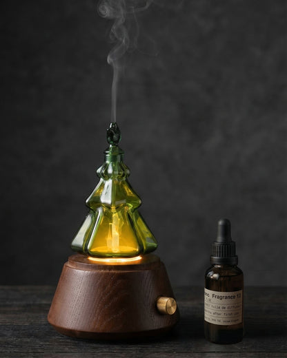 A detailed close-up product photograph of a specialized, multi-tiered clear glass waterless nebulizing diffuser shaped like a complex Christmas tree. The intricate internal glass capillary structure is glowing warmly from an integrated amber LED light, and a fine, cool mist is visibly curling upwards from the star-shaped top tip. The dark walnut wooden base plate features etched text 'Chavanc' and 'BEANCE AROMATHERAPY'. Placed on the aged wood surface next to the diffuser is an amber dropper bottle of 'BE.2