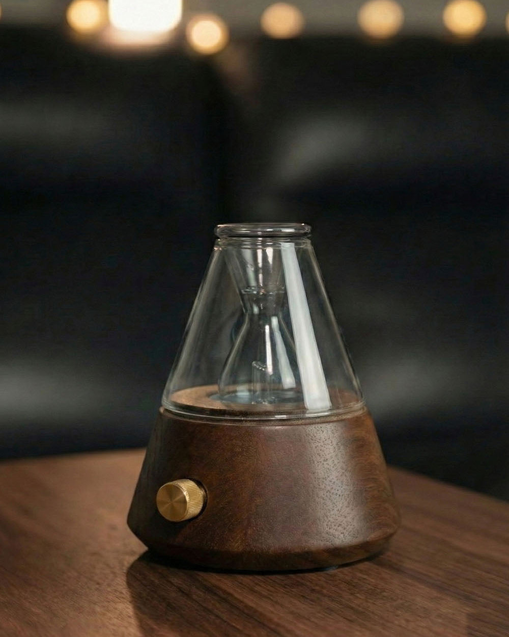 "A warm, detailed close-up of the Chavanc Aromawaterless nebulizing diffuser. The delicate hand-blown glass nebulizer is positioned next to a glowing Edison-style filament bulb, both mounted on a solid dark wood base with a knurled metallic dial. 