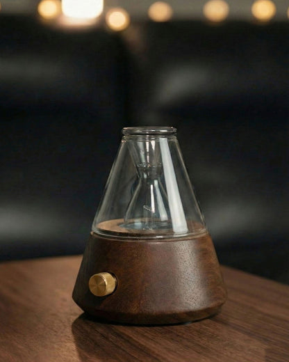 "A warm, detailed close-up of the Chavanc Aromawaterless nebulizing diffuser. The delicate hand-blown glass nebulizer is positioned next to a glowing Edison-style filament bulb, both mounted on a solid dark wood base with a knurled metallic dial. 