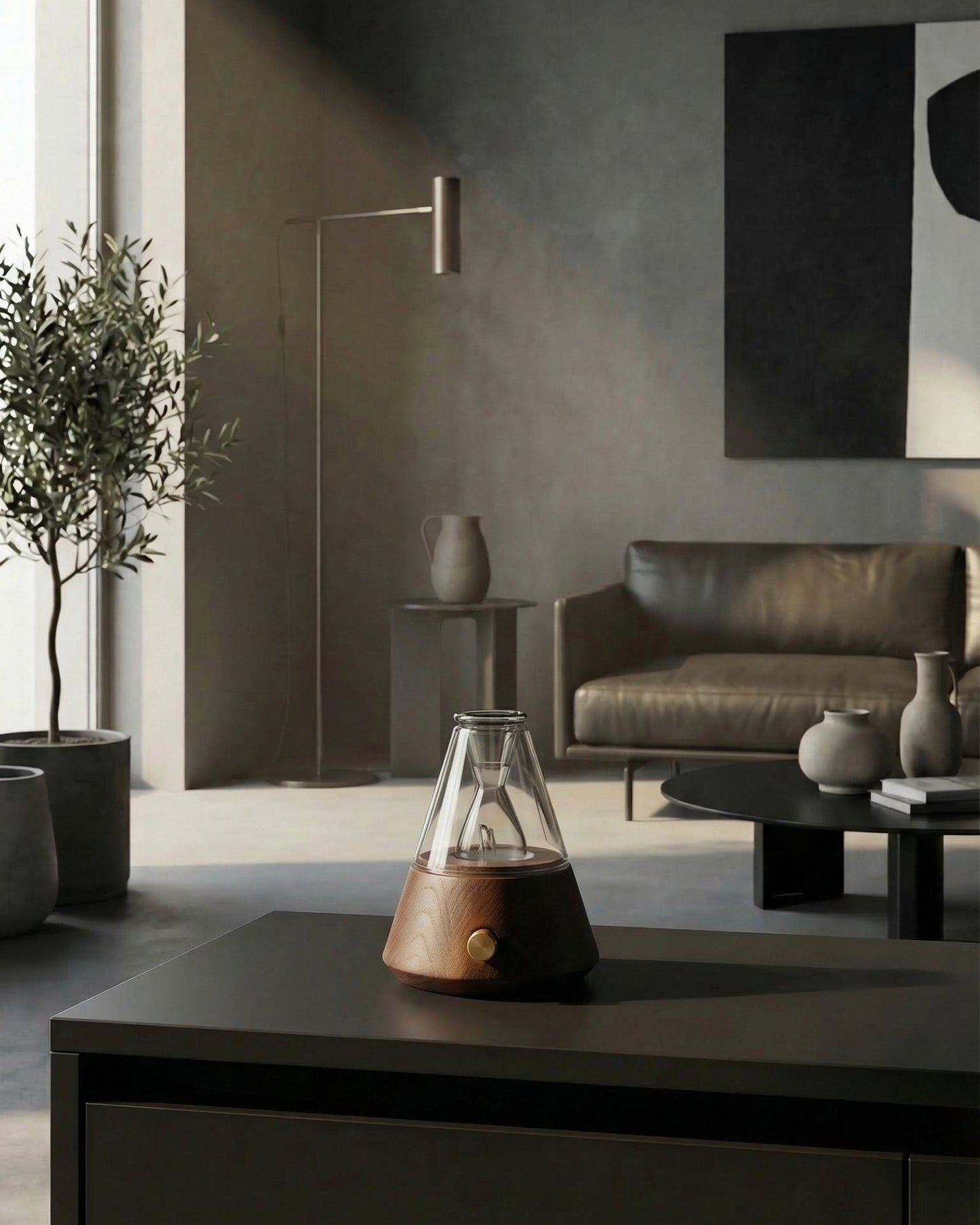 A high-end Chavanc Aroma waterless nebulizing essential oil diffuser with a tapered walnut wood base and a glass pyramid Nebula Flask. It is displayed on a dark console table in a sun-lit, modern living room featuring a concrete-grey wall, a tall olive tree, a dark leather sofa, and minimalist black-and-white wall art. The scene captures a sophisticated, high-end home decor and professional aromatherapy setup.