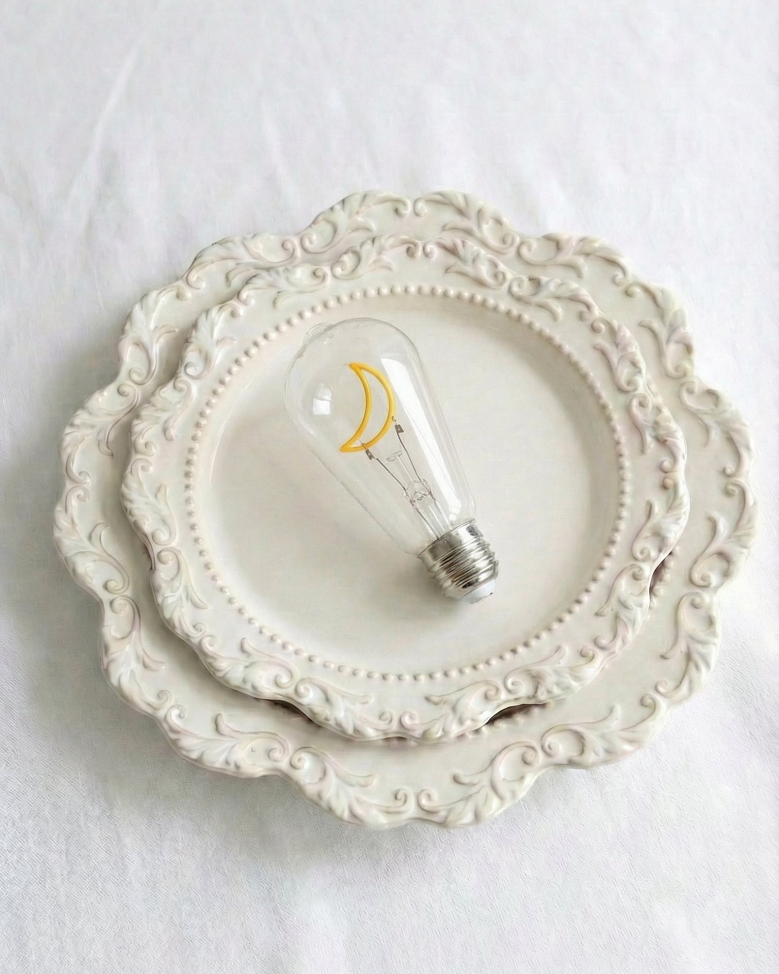 A detailed close-up photograph of a clear glass specialized replacement decorative bulb for a Chavanc_Araromatherapy diffuser, featuring an intricate, non-lighting, yellow moon-shaped decorative element (filament silhouette) inside. The metal connector base is visible at the bottom. The bulb rests on ornate, stacked cream-colored ceramic plates with scalloped edges and beaded borders. Perfect rustic aesthetic accessory for celestial home decor, high-end tablescapes, or home fragrance setup. conversation sta