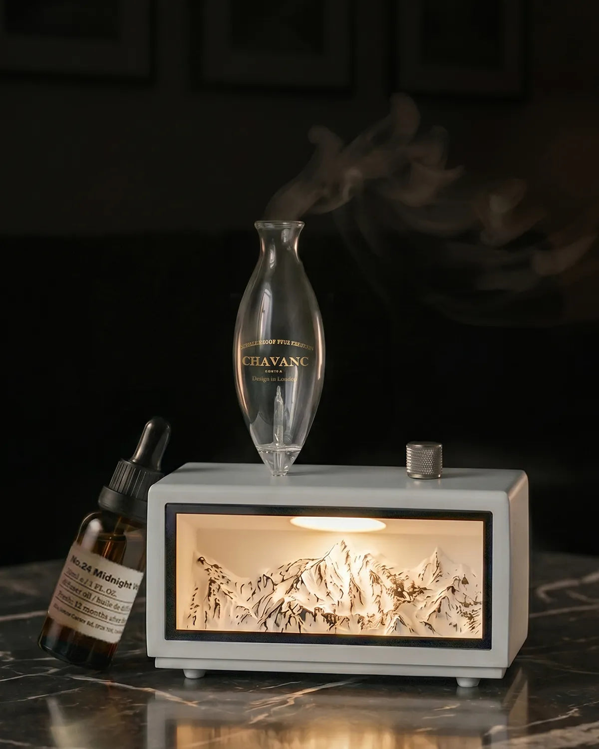 Chavanc Aroma waterless nebulizing diffuser featuring a backlit 3D alpine mountain relief inside a solid timber base, with a clear glass vessel emitting a plume of dry mist on marble.