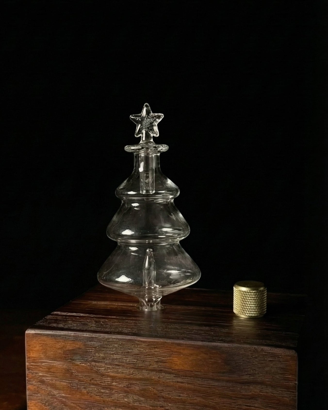 A sleek black rectangular Chavanc essential oil diffuser base featuring a clear glass Christmas tree-shaped nebulizing flask. The diffuser is emitting a delicate mist, placed next to a decorative jar of pinecones and dried flowers on a white cloth, creating a cozy winter holiday atmosphere.
