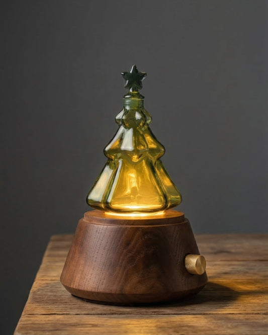 A professional studio product photograph of a Chavanc nebulizing aromatherapy diffuser. The device features a multi-tiered clear glass flask expertly shaped like a Christmas tree with a star on top. It sits on a premium round dark walnut wood base with a signature knurled gold control knob. The image is set against a dark, solid background to highlight the elegant, festive design and high-quality craftsmanship of the waterless essential oil diffuser.