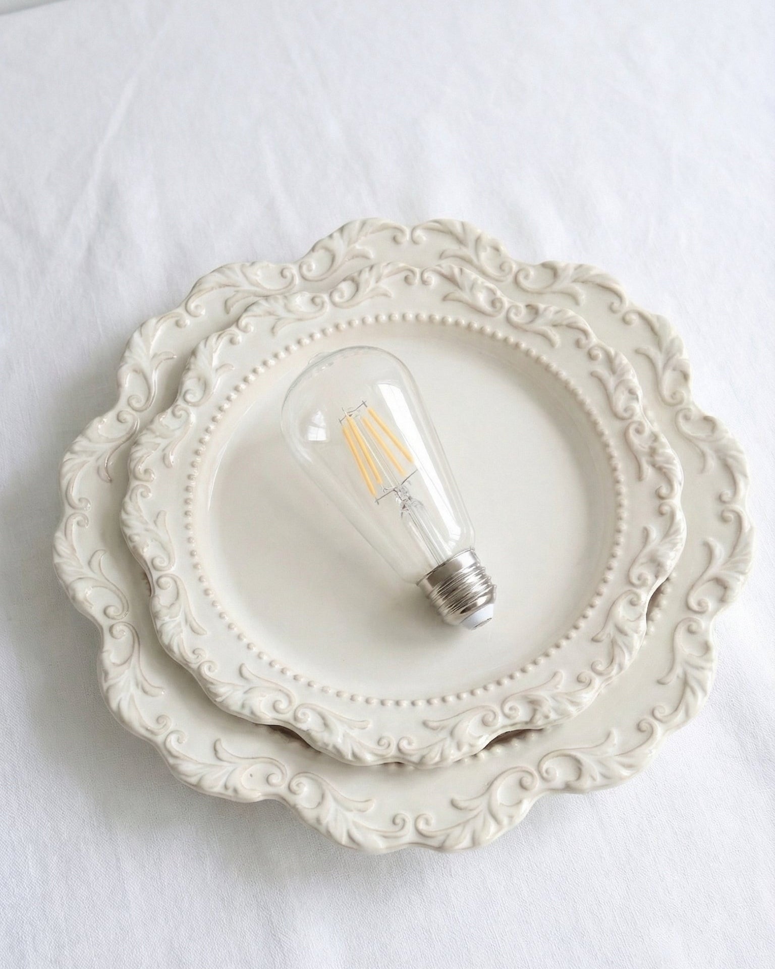 A classic ST64 clear glass LED replacement bulb for a Chavanc
aromatherapy diffuser, featuring four vertical warm-light filaments. The vintage Edison-style bulb is displayed on decorative, stacked cream ceramic plates with intricate scalloped edges, perfect for maintaining a timeless and elegant look for high-end home fragrance setups.
