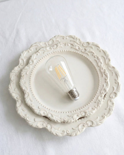 A classic ST64 clear glass LED replacement bulb for a Chavanc
aromatherapy diffuser, featuring four vertical warm-light filaments. The vintage Edison-style bulb is displayed on decorative, stacked cream ceramic plates with intricate scalloped edges, perfect for maintaining a timeless and elegant look for high-end home fragrance setups.