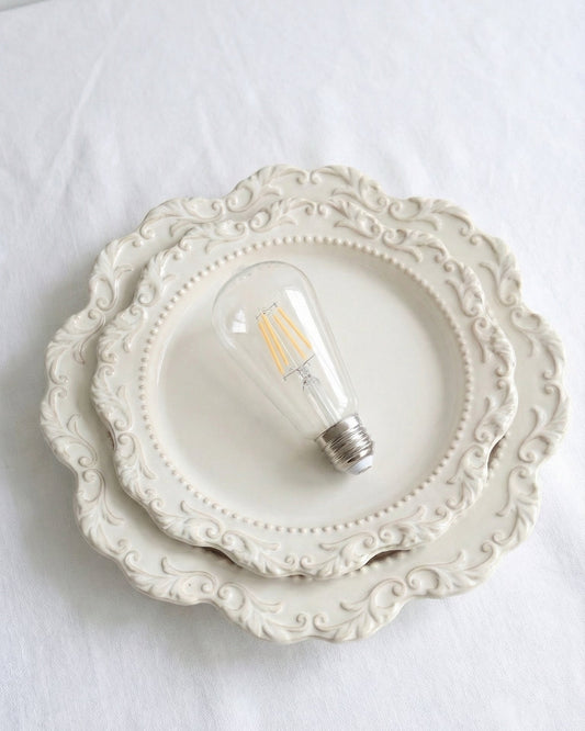A classic ST64 clear glass LED replacement bulb for a Chavanc
aromatherapy diffuser, featuring four vertical warm-light filaments. The vintage Edison-style bulb is displayed on decorative, stacked cream ceramic plates with intricate scalloped edges, perfect for maintaining a timeless and elegant look for high-end home fragrance setups.