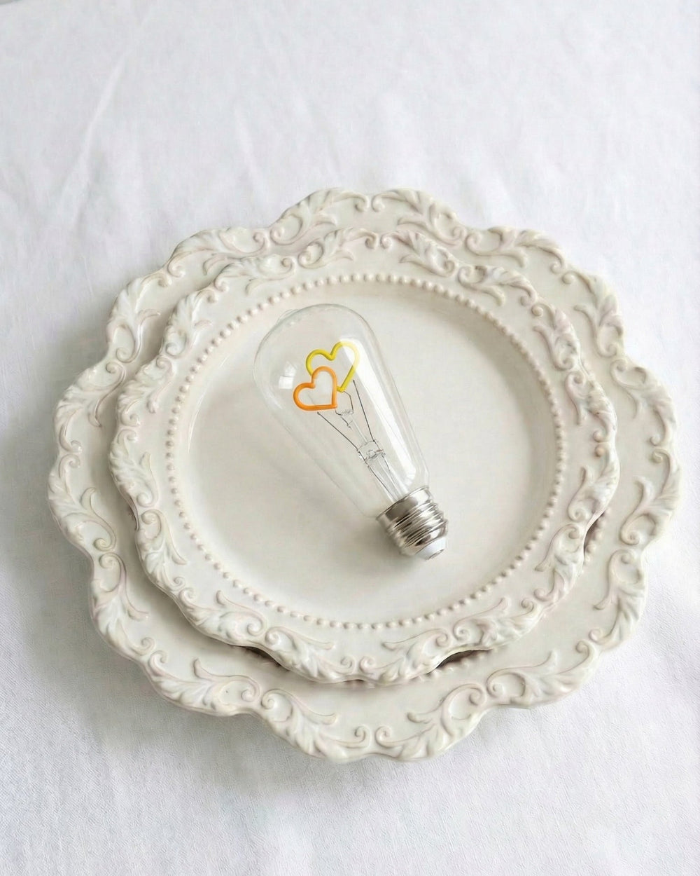 A specialized clear glass replacement bulb for a Chavanc aromatherapy diffuser, featuring two interlocking heart-shaped decorative filaments in yellow and orange. The vintage Edison-style bulb is displayed on ornate, stacked cream ceramic plates with scalloped edges, set against a white fabric background. A romantic and aesthetic home decor accessory for luxury diffusers.