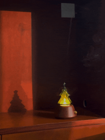 A close-up lifestyle photograph capturing a specialized clear glass replacement nebulizing flask for a Chavanc aromatherapy diffuser, shaped like a beautiful, tiered Christmas tree. The integrated yellow-orange iLED glows warmly from within, defining the tree's silhouette. A fine, cool mist is visibly curling from the star-shaped top. The flask rests on the signature stacked ornate, cream-colored ceramic plates with highly detailed scalloped edges and a beaded border, set against a white textured fabric b