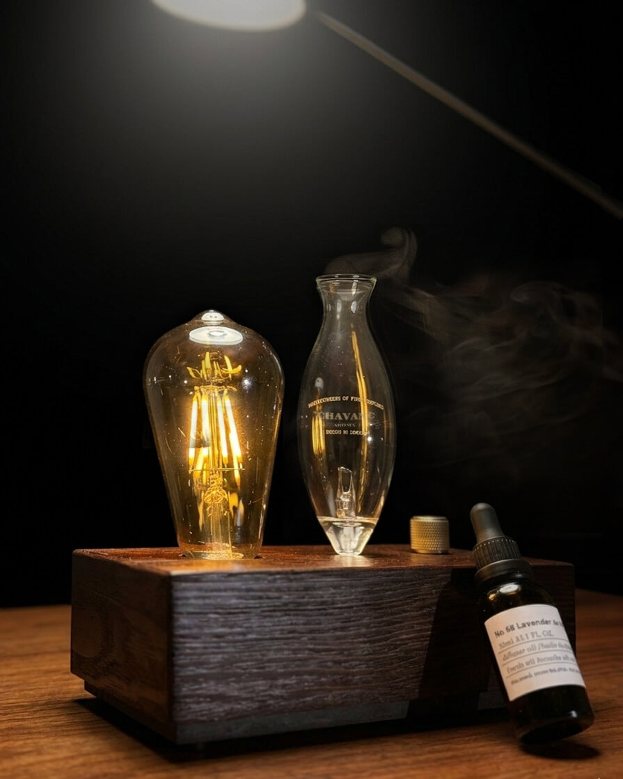 "A chavanc-Aroma waterless nebulizing diffuser featuring an illuminated Edison-style bulb that casts a warm amber glow. The instrument is crafted with a hand-blown glass nebulizer and a solid dark wood base, elegantly placed on a wooden table next to a signature amber essential oil bottle in a moody, sophisticated interior."