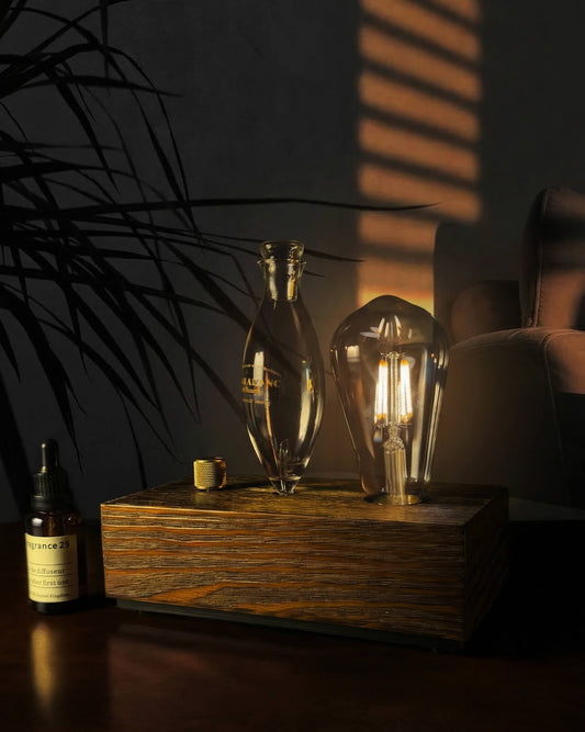 "A chavanc-Aroma waterless nebulizing diffuser featuring an illuminated Edison-style bulb that casts a warm amber glow. The instrument is crafted with a hand-blown glass nebulizer and a solid dark wood base, elegantly placed on a wooden table next to a signature amber essential oil bottle in a moody, sophisticated interior."