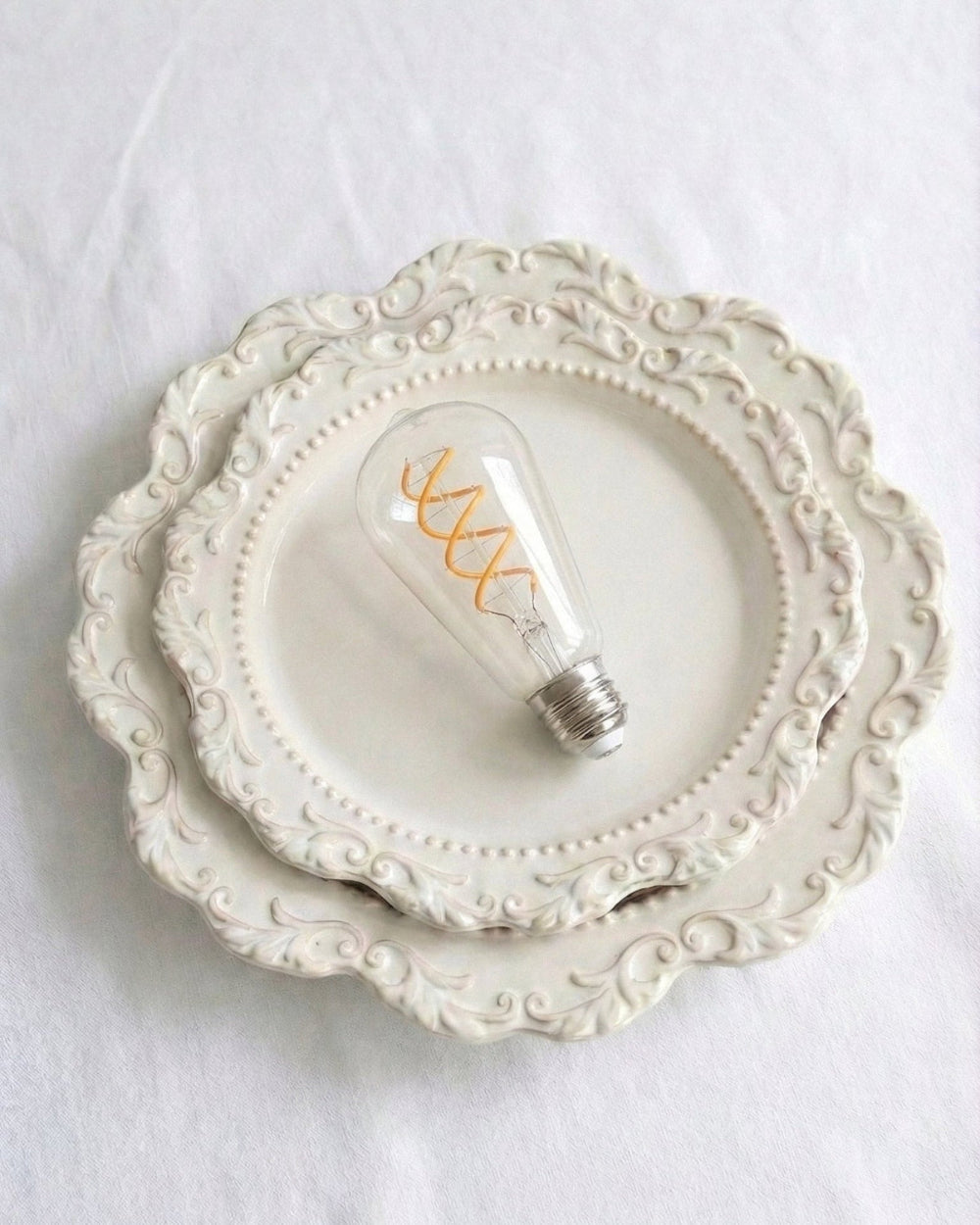 Detailed close-up photograph of a clear glass specialized replacement bulb for a chavanc- aroma aromatherapy diffuser, featuring a unique orange spiral helix-shaped decorative filament. The vintage Edison-style bulb rests on two ornate, stacked cream-colored ceramic plates with scalloped edges and beaded borders. An aesthetic and retro accessory for home fragrance and luxury interior decor.