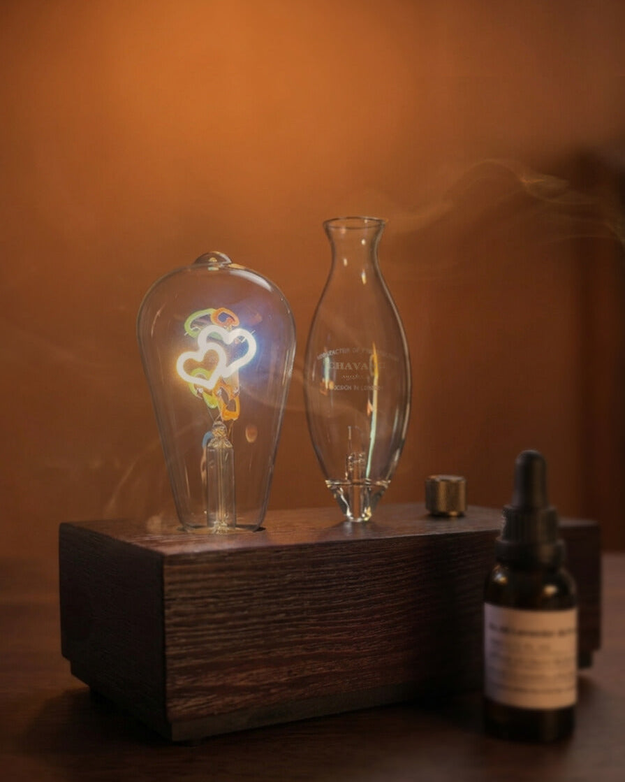 "A warm, detailed close-up of the chavanc-aroma waterless nebulizing diffuser. The delicate hand-blown glass nebulizer is positioned next to a glowing Edison-style filament bulb, both mounted on a solid dark wood base with a knurled metallic dial. A bottle of chavanc-aroma 29 sabtal essential oil sits beside it, highlighting the artisanal quality."