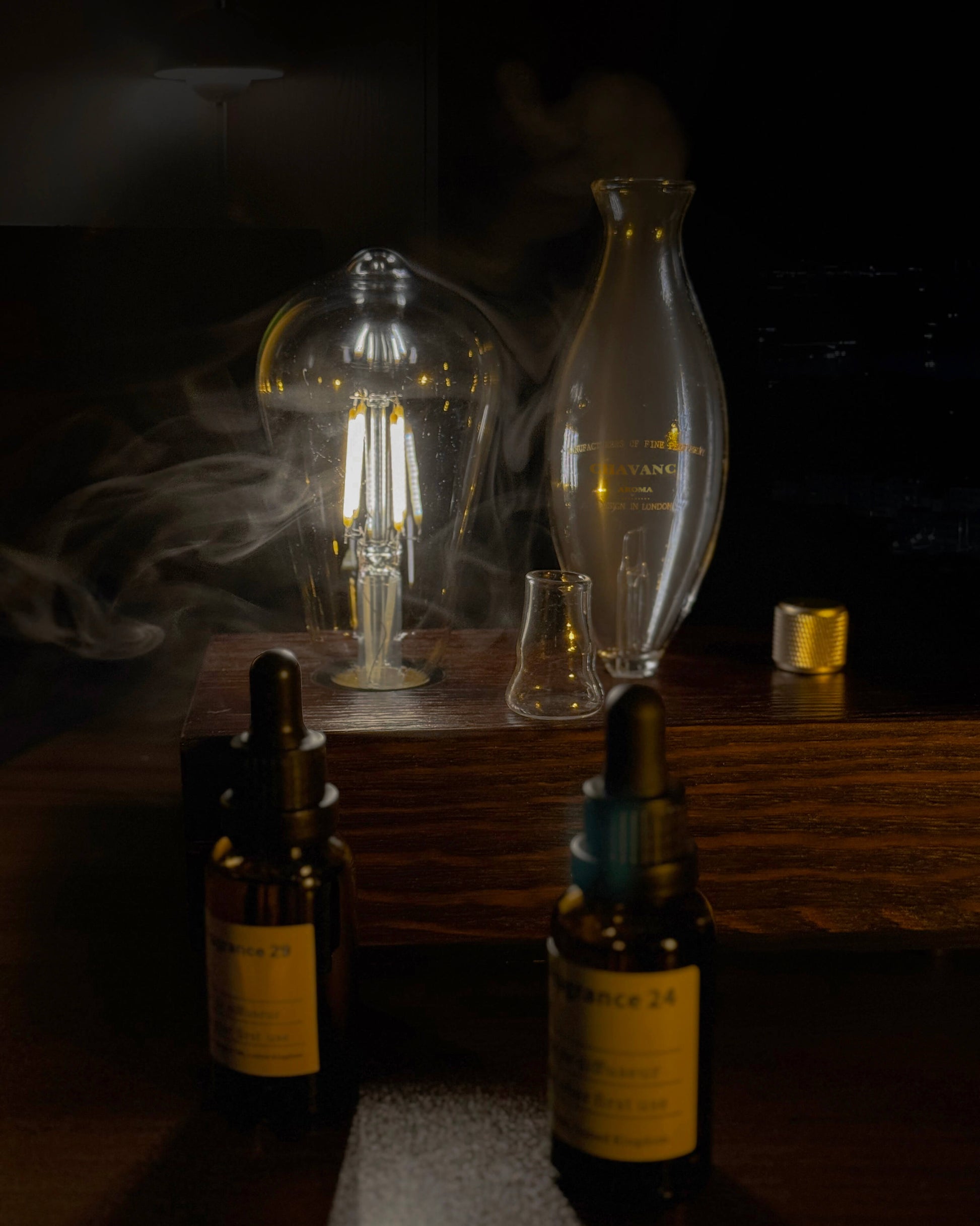 "A warm, detailed close-up of the chavanc-aroma waterless nebulizing diffuser. The delicate hand-blown glass nebulizer is positioned next to a glowing Edison-style filament bulb, both mounted on a solid dark wood base with a knurled metallic dial. A bottle of chavanc-aroma 29 sabtal essential oil sits beside it, highlighting the artisanal quality."