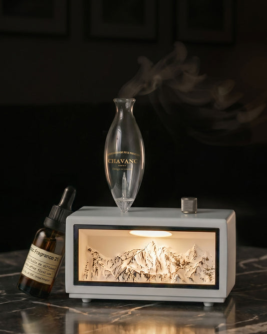 Chavanc Aroma waterless nebulizing diffuser featuring a backlit 3D alpine mountain relief inside a solid timber base, with a clear glass vessel emitting a plume of dry mist on marble.