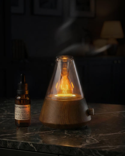 Chavanc Aroma conical waterless nebulizing diffuser with solid timber base and cone-shaped glass housing a warm glowing ambient lamp, emitting dry mist on a marble table.