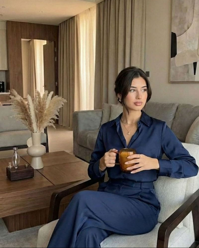 "A sophisticated lifestyle shot in a high-end modern apartment. A woman in a navy blue silk blouse and matching trousers sits elegantly on a light grey armchair, holding a glass of coffee. In the background, the chavanc-aroma waterless scent diffuser with a walnut wood base is placed on a dark wood coffee table next to a minimalist pampas grass arrangement, blending professional style with domestic serenity."