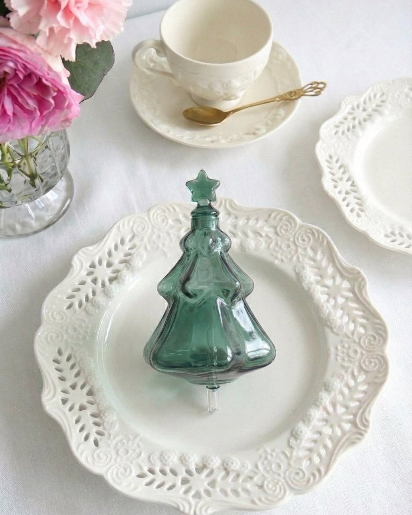 A beautiful emerald green tinted glass nebulizing flask in the shape of a Christmas tree, designed as a replacement accessory for Chavanc essential oil diffusers. The festive green glass features a tiered tree design and rests on ornate, stacked cream-colored ceramic plates with vintage scalloped edges. Fine aromatherapy mist is visible curling from the star-shaped top. Perfect for forest-inspired home decor and holiday seasonal fragrance setups.