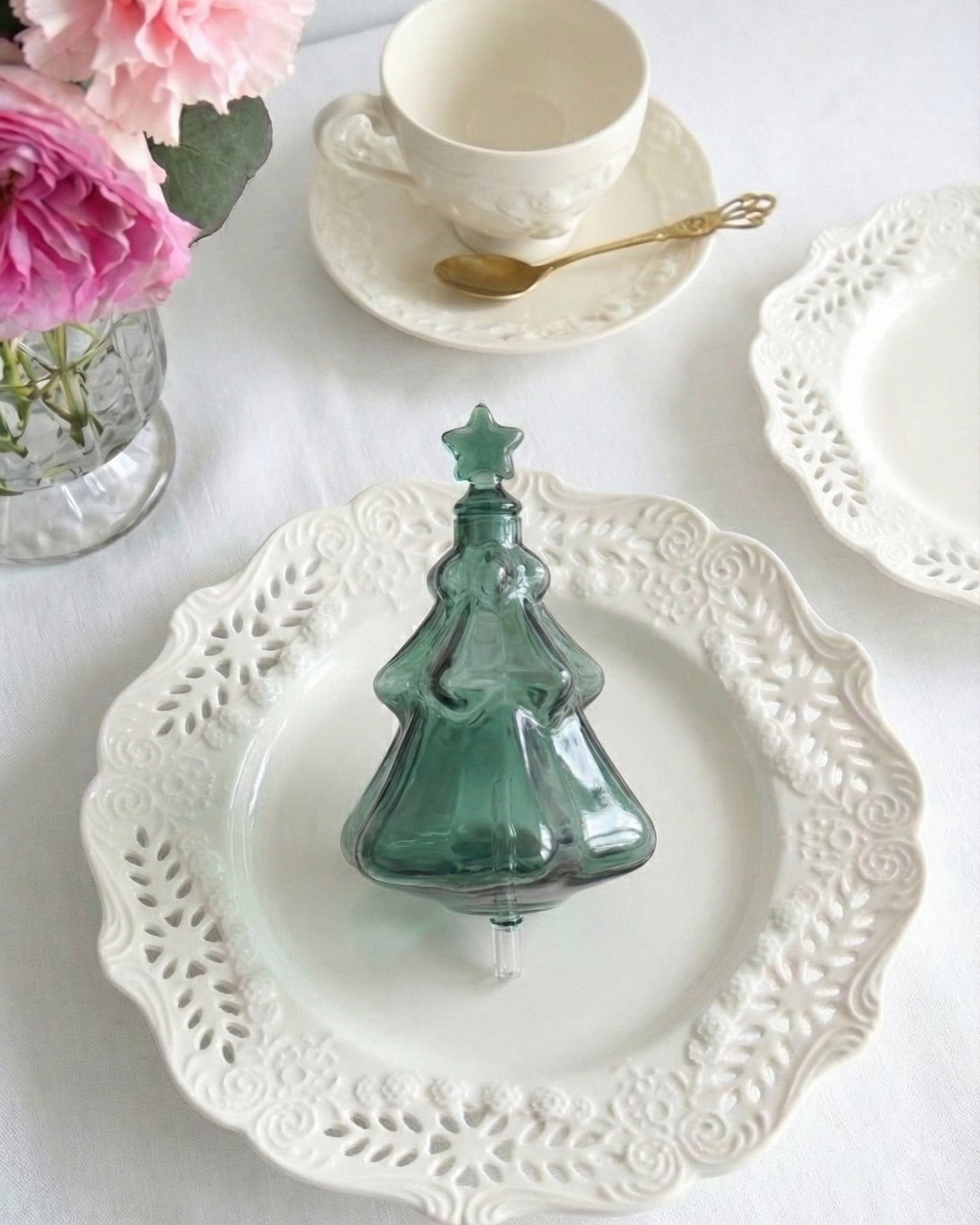 A beautiful emerald green tinted glass nebulizing flask in the shape of a Christmas tree, designed as a replacement accessory for Chavanc essential oil diffusers. The festive green glass features a tiered tree design and rests on ornate, stacked cream-colored ceramic plates with vintage scalloped edges. Fine aromatherapy mist is visible curling from the star-shaped top. Perfect for forest-inspired home decor and holiday seasonal fragrance setups.
