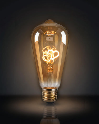 A specialized clear glass replacement bulb for a Chavanc aromatherapy diffuser, featuring two interlocking heart-shaped decorative filaments in yellow and orange. The vintage Edison-style bulb is displayed on ornate, stacked cream ceramic plates with scalloped edges, set against a white fabric background. A romantic and aesthetic home decor accessory for luxury diffusers.