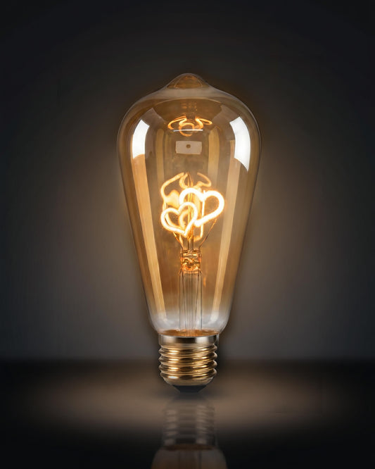 A specialized clear glass replacement bulb for a Chavanc aromatherapy diffuser, featuring two interlocking heart-shaped decorative filaments in yellow and orange. The vintage Edison-style bulb is displayed on ornate, stacked cream ceramic plates with scalloped edges, set against a white fabric background. A romantic and aesthetic home decor accessory for luxury diffusers.